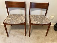 Front view of two teak dining chairs with fabric seats against a plain wall, showing the overall condition and color of the upholstery and wood finish.