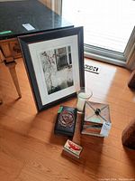 Framed print, two boxes, glass candle holder with candle, and match box shown on wooden floor near window.