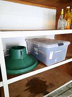 Photo of green plastic Christmas tree stand and clear plastic bin with Christmas lights on wooden shelf.