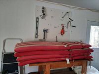 Six stacked lounge chair cushions on a wooden table in a garage workspace, red fabric with beige and white stripes visible on sides