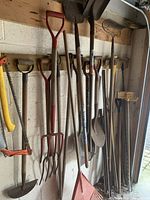 Photo showing an array of garden tools hanging on a wall including shovels, rakes, pitchforks, and hand saws, all with wooden handles and showing signs of wear.