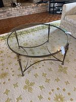 Full view of round glass coffee table on carpet showing glass top and brass metal base with four legs and curved support.