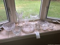Overview of eight clear glass items on window ledge showing bowls, vases, pitchers, and compote dish.
