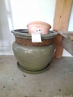 Photo showing large green glazed ceramic planter with smaller terracotta pot inside, placed on concrete floor.