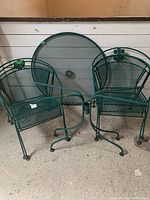 The image shows two green metal mesh chairs and the round table with a mesh top and umbrella hole, placed indoors on a concrete floor.