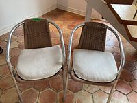 Two petit outdoor chairs with silver tubular metal frames, brown woven backs and seats, each with off-white removable cushions placed on the seats. Shown indoors on hexagonal tile flooring.