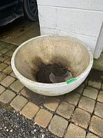 Concrete round planter placed outside near a garage with a brick-paved floor. Shows wear and dirt inside and outside, indicating previous use.