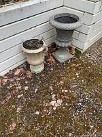 Two outdoor planters placed on mossy ground near white wooden wall. One shorter, plain design; other taller with rope pattern.