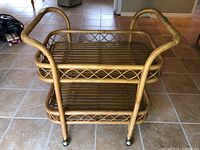Front and side view of a solid rattan serving trolley with two slatted shelves, missing top glass shelf, curved handles, and caster wheels on tile floor.