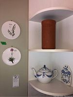 Shelved ceramic items: Royal Copenhagen blue and white teapot, tall rustic clay wine cooler, floral ceramic wall plate leaning beside teapot, and two botanical decorative plates hanging on the nearby wall.
