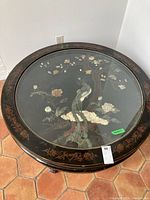 Full view of round black lacquer coffee table with ornamental Asian-inspired painted birds and flowers under glass top.