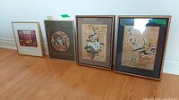 Photo of four framed wall art pieces including two Chinoiserie bird scenes and one European village landscape, arranged side by side on a wooden floor.