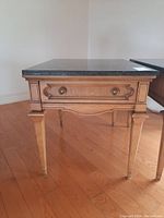 Wood side table front view showing drawer with brass handles and decorative wood details under a black granite top.