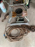 Black and Decker Workmate folding tool work bench with wooden top panels showing wear. Various tools and accessories placed on and around the bench including extension cord, pulley, axe head, wedges, gloves, container with screws, and black bucket.