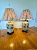 Pair of large Chinoiserie style porcelain table lamps with pleated fabric shades placed on hardwood floor, lit up showing warm light diffusion. Each lamp features floral and bird decorations on white porcelain and dark wooden bases.