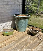 Three plant pots on a wooden deck alongside a brick wall, including one large green round pot, one medium round pot with dried plants, and one small rectangular green pot with soil residue.