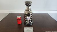 14 inch silver-colored Stanley Cup replica with engraved band plates, autographed by Marcel Dionne, next to a Coca-Cola can for size reference.