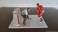 Side angle showing Gordie Howe in red facing off against goalie Terry Sawchuk in blue, standing on rectangular rink base with net behind Sawchuk.