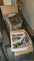 Photo showing a wire basket filled with various printed automotive manuals, some vintage, along with stacks of Muscle Machines magazines on top of a cardboard box.