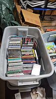 Plastic bin filled with CD jewel cases in a garage setting, showing stacked rows of assorted music CDs.