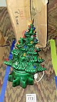 Photo showing green ceramic Christmas tree with colorful bulbs and yellow star topper, with visible power cord, against a patterned background and adjacent cardboard box.