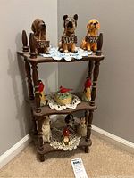 Three-tier dark wood display shelf with various ceramic dog and bird figurines arranged on it in corner against wall.