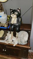 Black and white cat figurine standing upright, showing wear and scuffs, with two Beanie Babies and a stone bear on a wooden surface.