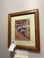 Framed racoon art print hung on a wall, showing the full piece including wooden frame and matting, with a maxsold tag.