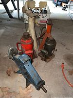 Photo showing all the jacks and jack stands in the lot including the blue scissor jack, 2 red bottle jacks, black bottle jack, and 2 jack stands (orange and white) with visible dirt and rust.