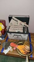 Metallica Live Shit VHS box set with multiple tapes arranged inside alongside brass bugle and wooden paddle board with flame artwork on a wooden surface.