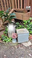 Overall view of the milk can, metal box, and small planter with faux flowers placed outdoors on ground with ivy background.