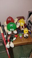 Photo showing five M&M character items: large red, green, yellow dispensers with expressive faces, a small green dispenser shaped like a car, and a brown dispenser with racing attire, all on wooden table surface.