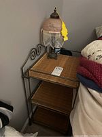 Metal and wicker three-tier side table with note on top shelf next to vintage Victorian style lamp with tasseled frosted glass beaded shade, showing fringe partially detached.