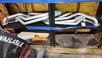 Long view of metallic small block Chevy headers laid on a shelf in a garage, showing multiple tubular pipes converging.