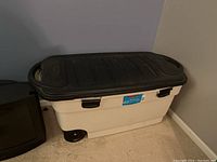 Storage tub with black lid placed on carpet floor against a wall.