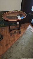 Full view of round wood side table with scenic image under glass top, standing on wooden floor near wall and rug.