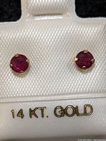 Close-up image of pair ruby stud earrings mounted in 14K yellow gold prong settings, displayed on a white card labeled '14 KT. GOLD'. The stones are round and bright red.