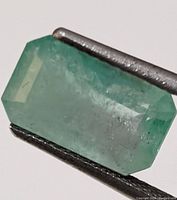 Close-up of the natural emerald gemstone held with tweezers showing the faceting and internal inclusions.
