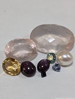 Photo showing nine mixed gemstones of various sizes and colors including pale pink, yellow, purple, teal blue, blue, and one pearl
