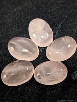 Five oval polished rose quartz stones displayed on black fabric, showing their translucent pale pink color and internal inclusions.