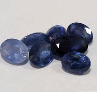 Six genuine sapphire gemstones in a range of blue shades and oval shapes on a plain light surface. The stones display faceting and natural variation in color and clarity.