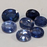 Seven oval-cut blue sapphire gemstones arranged on a white surface, showing varying blue tones and faceted cuts.