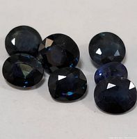 Seven deep blue faceted sapphire gemstones shown scattered on a neutral surface. Stones have oval and round shapes, displaying natural color variation and internal inclusions.
