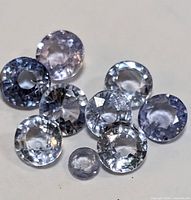 Photo showing ten loose round faceted tanzanite gemstones in various light violet-blue colors and sizes, arranged closely together on a neutral background.