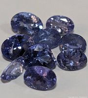 Photo showing eleven faceted oval tanzanite gemstones in shades of violet-blue and purple with visible inclusions and some damage on stones.