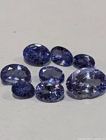 Close up of nine loose oval tanzanite gemstones in varied blue/violet hues and clarity, some showing internal inclusions and minor breakage.