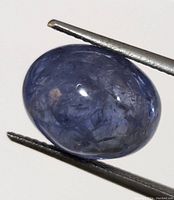 Close-up of oval cabochon cut tanzanite gemstone held by tweezers showing blue-violet color and internal inclusions.