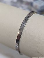 Close-up photo of a silver bracelet with an angular segmented pattern on a white curved display surface.