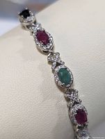 Close-up image showing alternating oval ruby, emerald, and sapphire stones set in sterling silver with small clear stone accents.
