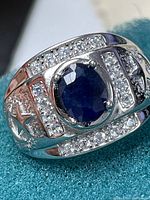 Close-up photo of men's silver ring with central blue sapphire and surrounding cubic zirconia stones showing star-shaped band cutouts.